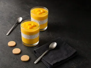 Dois copinhos de smoothie de manga e chia em camadas ao lado de três fichas de madeira sem marcação