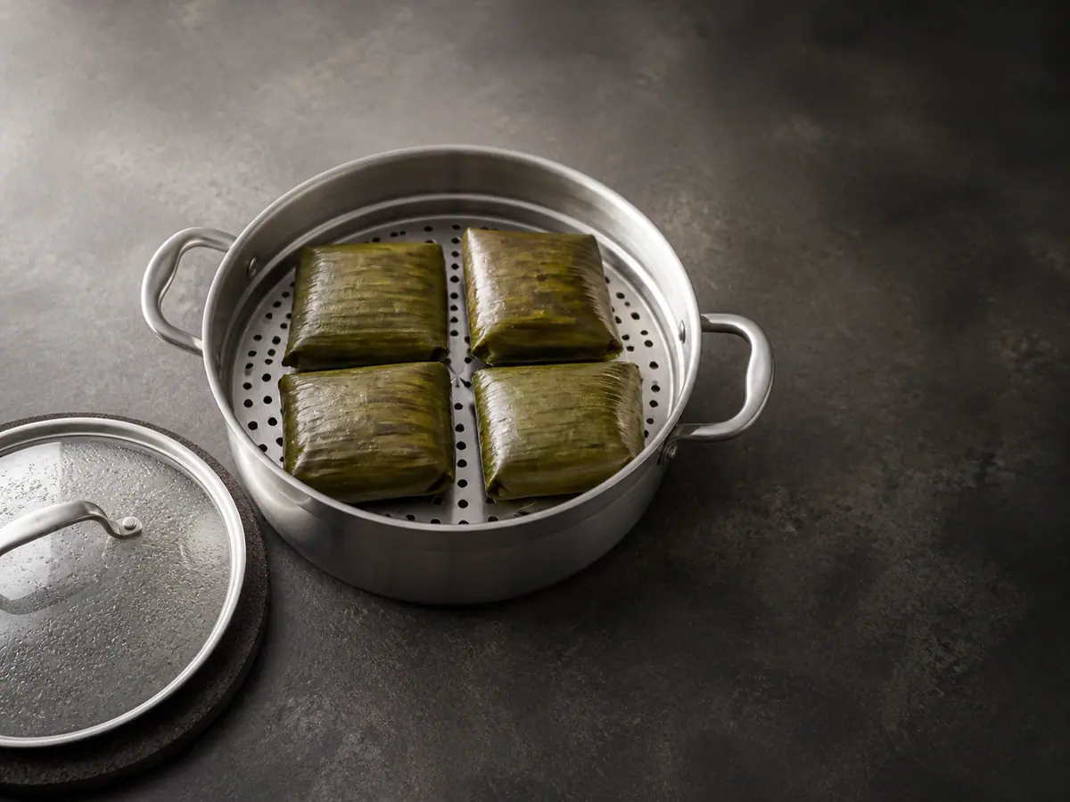 Four deep-olive steamed banana-leaf parcels spaced in a steamer basket