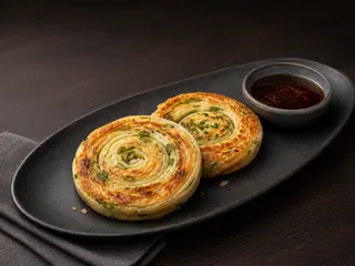 Duas espirais douradas de panqueca de cebolinha em sequência ao lado de molho de shoyu com gengibre