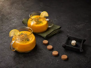 Two flask-shaped mugs of golden lemon lentil soup with parsley, lemon wedges, turn markers, and a die