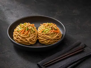 Dos nidos de fideos con maní y sésamo, pepino, zanahoria, cebollín, semillas de sésamo y maní en un tazón oscuro y poco profundo