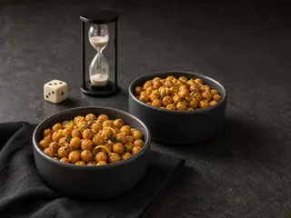 Dos vasitos de garbanzos tostados y crujientes con romero y limón junto a un dado y un pequeño reloj de arena