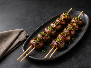 Dois espetinhos duplos de cogumelos teriyaki cobertos com molho, dispostos em uma curva com cebolinha e gergelim