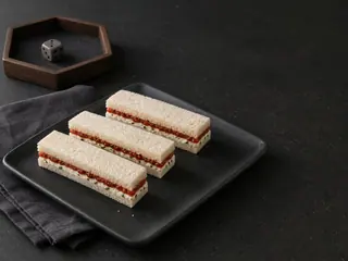 Cuatro tiras de sándwich de té sin corteza con mermelada de tomate y queso crema con cebollín, sobre un plato oscuro junto a un dado sencillo en su bandeja