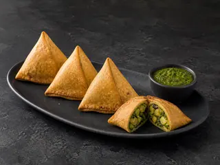 Four baked spinach samosa pockets with green chutney, including one opened pocket showing the potato, pea, and spinach filling