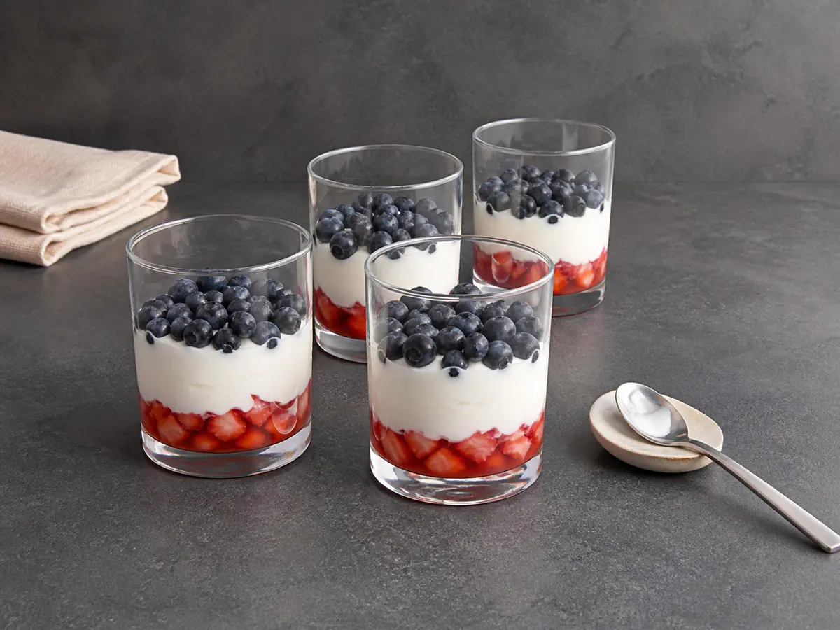 Four parfait glasses layered with strawberry mixture, Greek yogurt, and blueberries before granola